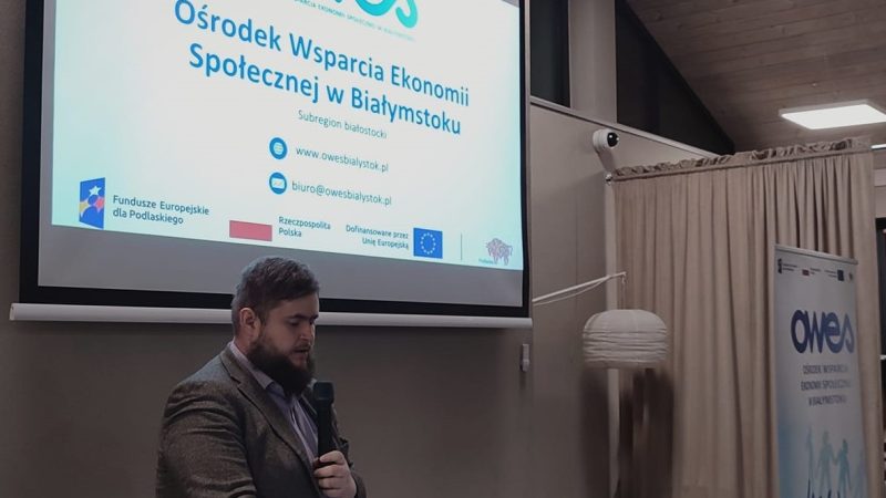 Mężczyzna z brodą mówi do mikrofonu. W tle prezentacja dotycząca OWES wyświetlona na ekranie.