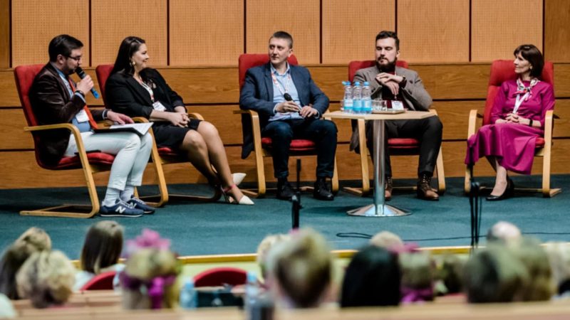 Pięć osób, kobiet i mężczyzn siedzi na krzesłach na scenie. Przygląda im się ludzie siedzący na widowni.